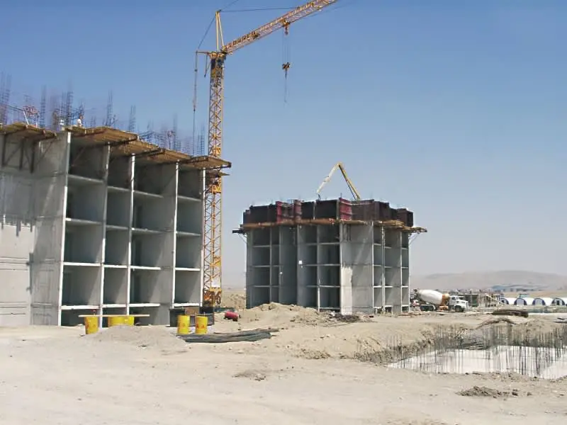 Kurak şantiye alanında betonarme yapı çekirdeği inşaatı; solda tünel kalıbıyla inşa edilmiş hücre duvar sistemi tamamlanmış beyaz beton, sağda yuvarlak kesitli başka bir beton silo yapısı ve üstünde inşaat vinci görülmekte