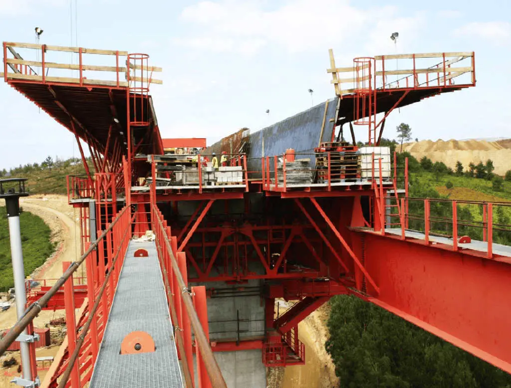 Yakın plan MSS köprü ayağı bağlantısı; kırmızı çelik kafes sistem pier üzerine oturmuş, üst platformlarda çalışma alanları ve akıllı kilitleme mekanizmaları görülmekte