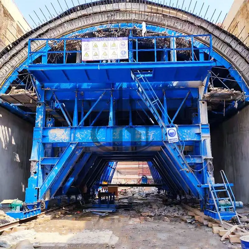 Tünel içinde mavi çelik tünel kalıp arabası; giriş açıklığında arkasında kazılmış kaya yüzeyi ve üst kısımda çimento sızıntısı ile uyarı levhaları, önde çalışanlar için merdivenler görülmekte