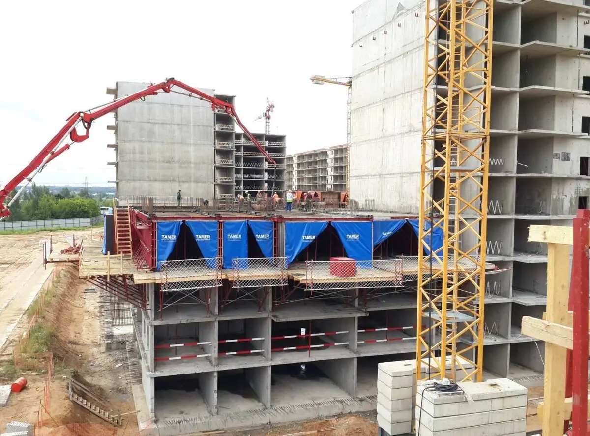 Şantiye havadan görünümü: kırmızı beton pompası kollu vinç, çok katlı bina iskeleti ve ön planda tünel kalıbı modülleri; çevrede diğer bina iskeletleri ve inşaat vinçleri görülmekte
