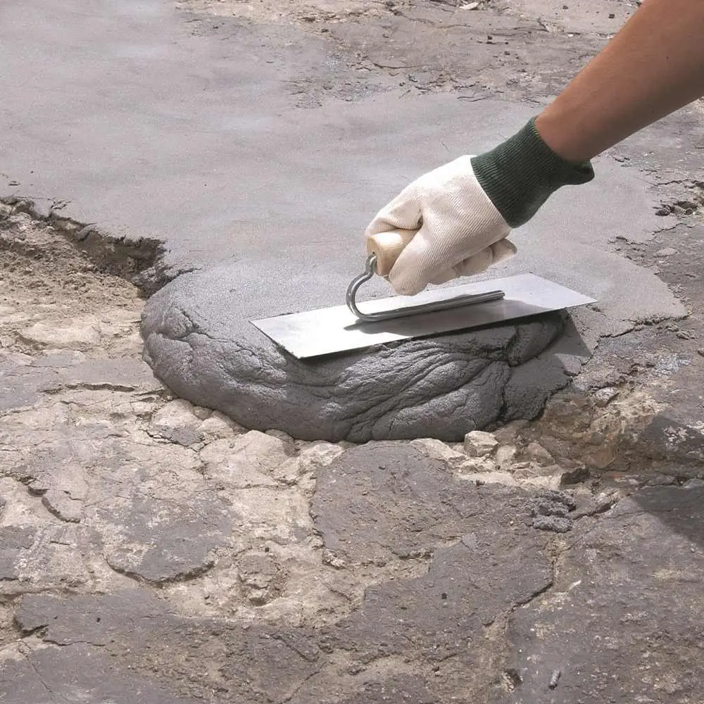 Beton yüzey onarımı yakın plan; bir el yeşil koruyucu eldiven ile spatula benzeri alete benzer küçük bir aletle büyük bir beton kırık/boşluk üzerine gri çimento tamir harcı sürüyor