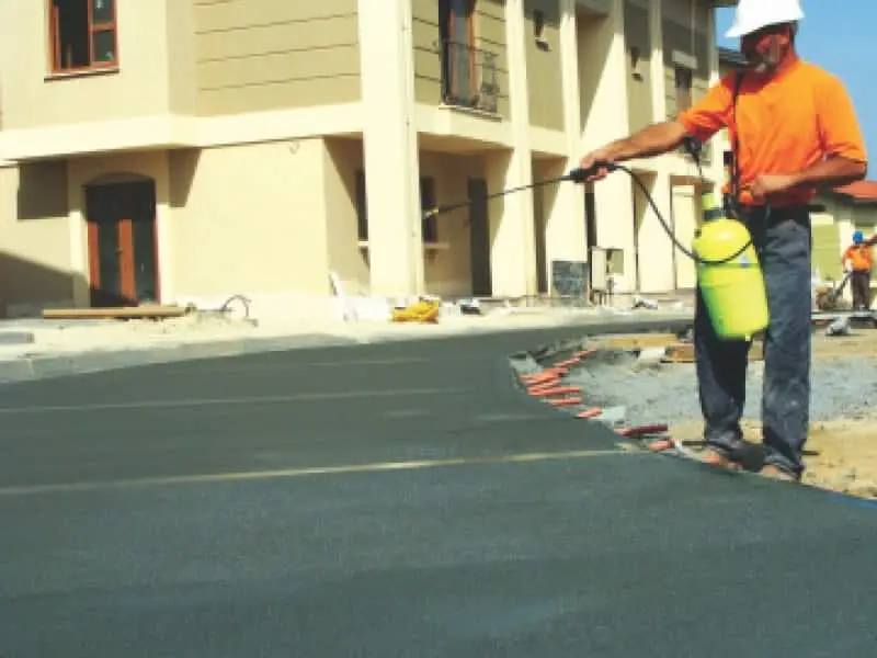 İşçi taze beton döşemeye kür spreyi uyguluyor — turuncu yelekli teknisyen arka planda bina ile