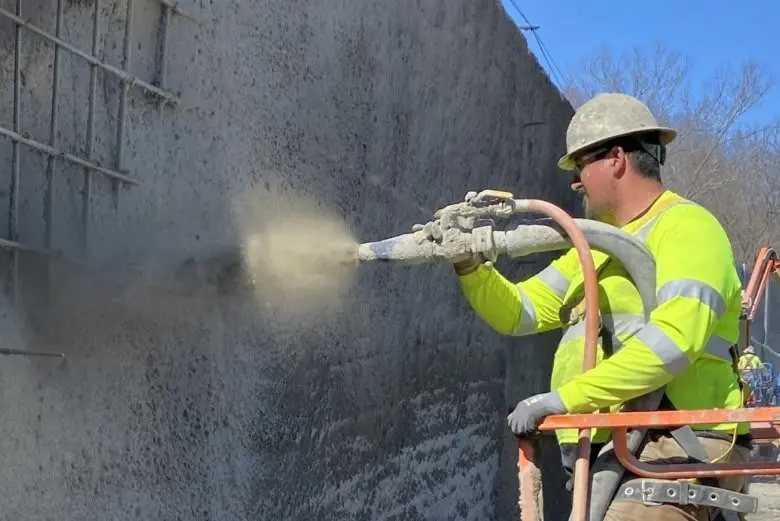 İşçi shotkrit (püskürtme beton) uyguluyor — sarı yelekli teknisyen yüksek basınçlı nozulla betonarme duvara