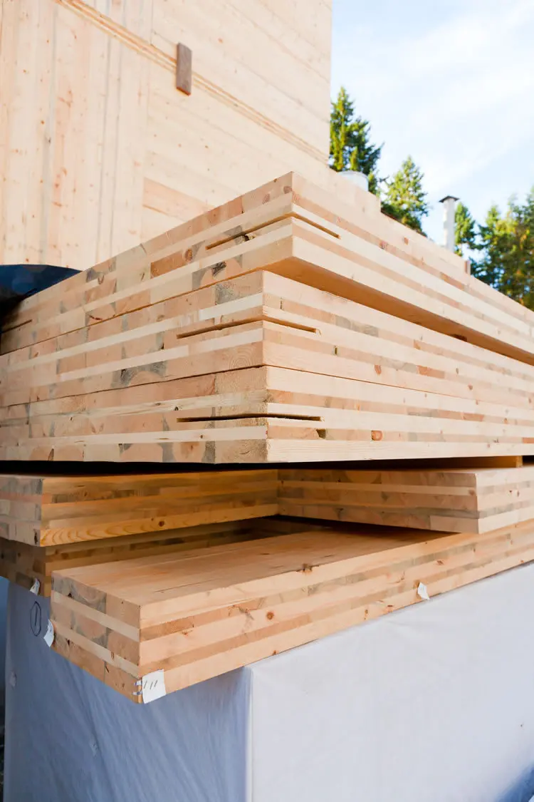 Açık alanda istiflenmiş CLT/glulam panelleri — etiketli, katmanlı ahşap levhalar ve arka planda bina