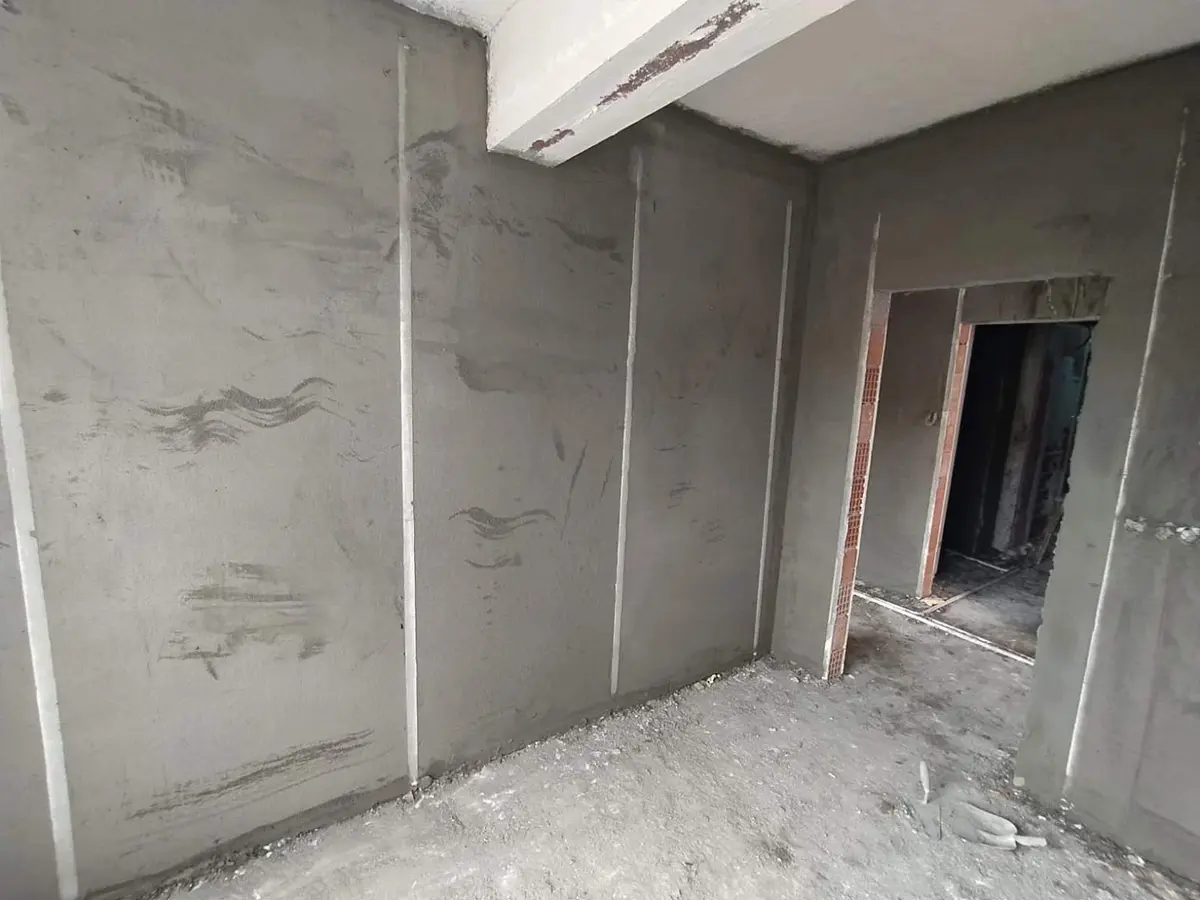 İnşaat halindeki dairenin iç mekanı; beton kolon izleri görünen gri çimento sıvalı duvarlar, kiriş altında sıva derz çizgileri, açık kapı boşluğu ve bims/tuğla geçiş yüzeyi