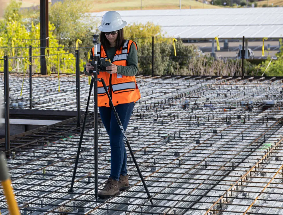 Şantiyede betonarme temel plağı donatı kafesi üzerinde turuncu yelek ve baret giyen kontrol mühendisi total station ile kesin hesap için metraj ölçümü yapıyor