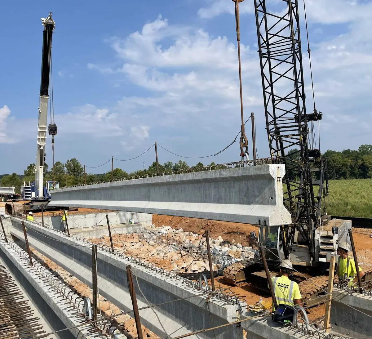 Köprü inşaatında vinç yardımıyla kaldırılan büyük öngerilmeli prefabrik beton I-kirişleri — sahada montaj aşaması, işçiler betonarme ayaklar üzerinde konumlandırıyor