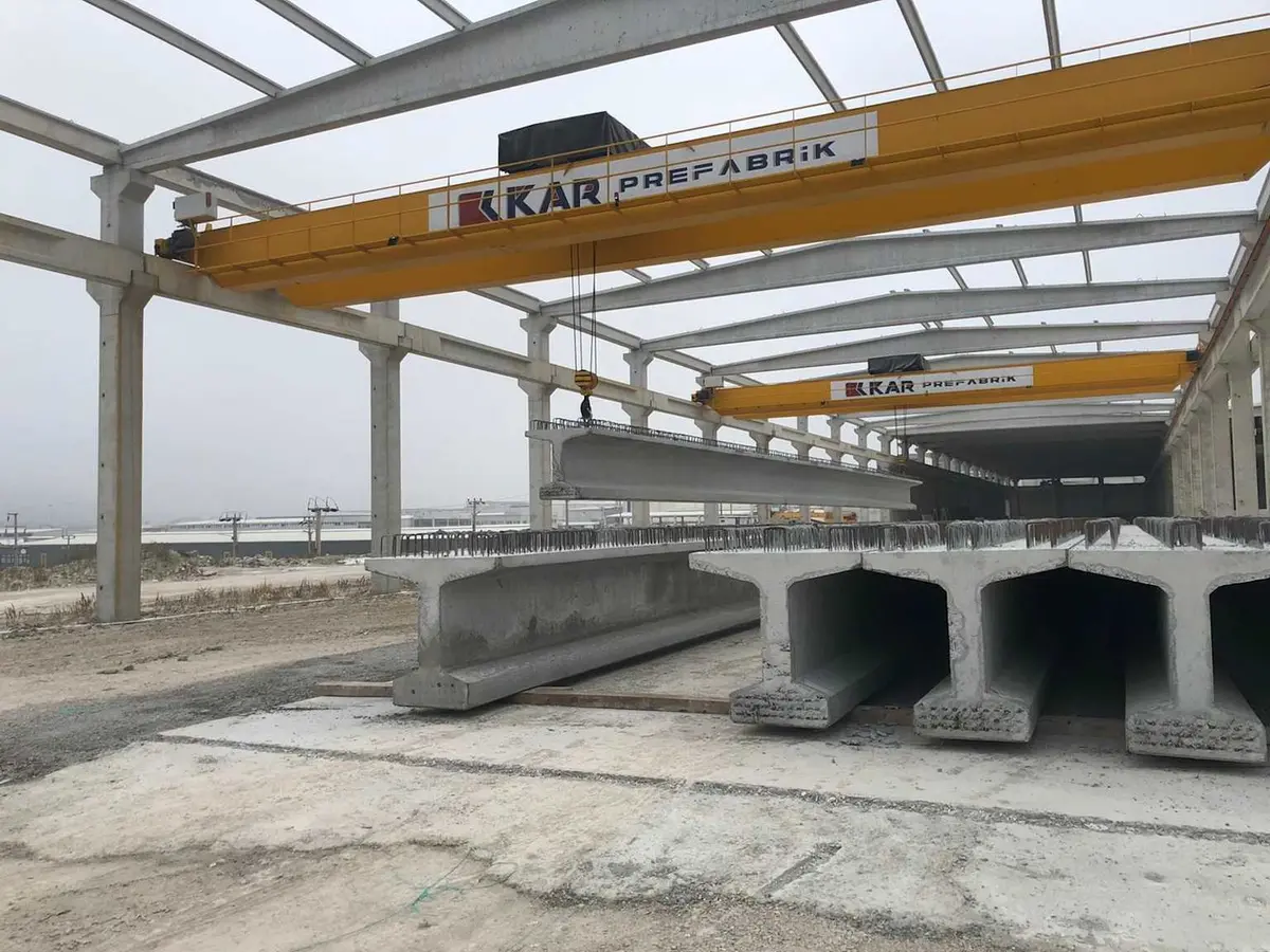 KAR Prefabrik fabrikasında köprü kirişlerinin köprü vinciyle kaldırılması — büyük açıklıklı I-kiriş yatay konumda askıda