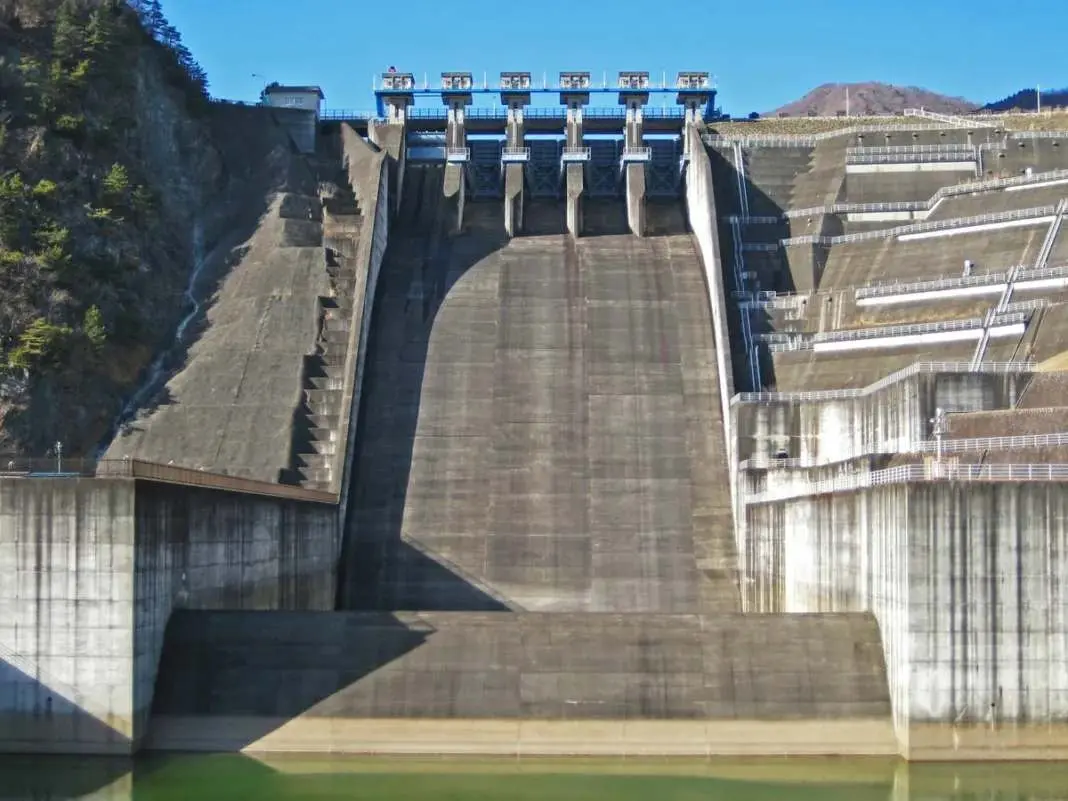 Büyük beton baraj savağı (spillway/ogee) mansap görünümü — açık kapılar, eğimli ogee yüz profili ve mansap dönüm yapısı