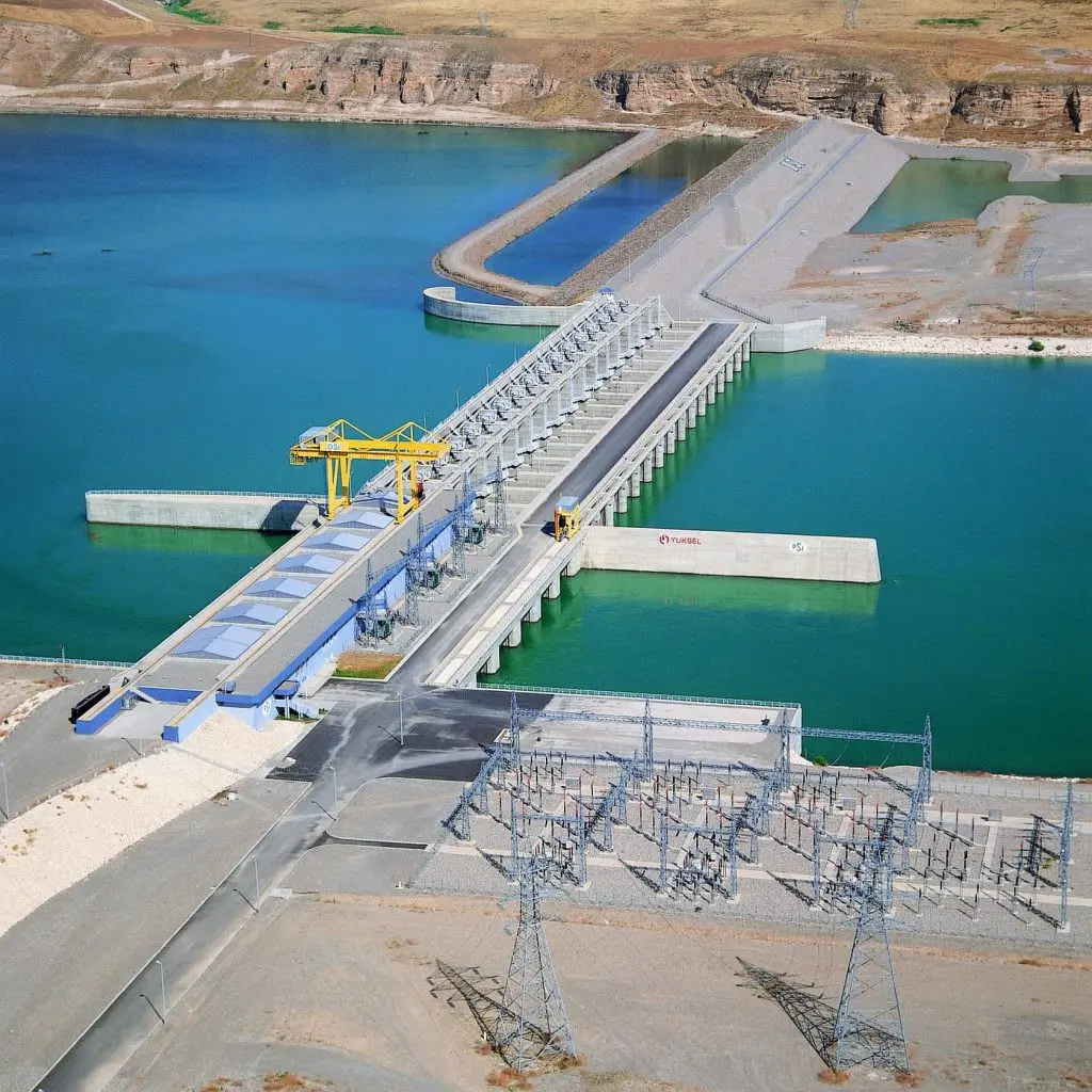 Büyük sulama ve enerji barajı havadan fotoğrafı — düz tip beton baraj gövdesi, açık kanal tahliye yapısı, sarı gantry vinç ve enerji santralı iletim hatları
