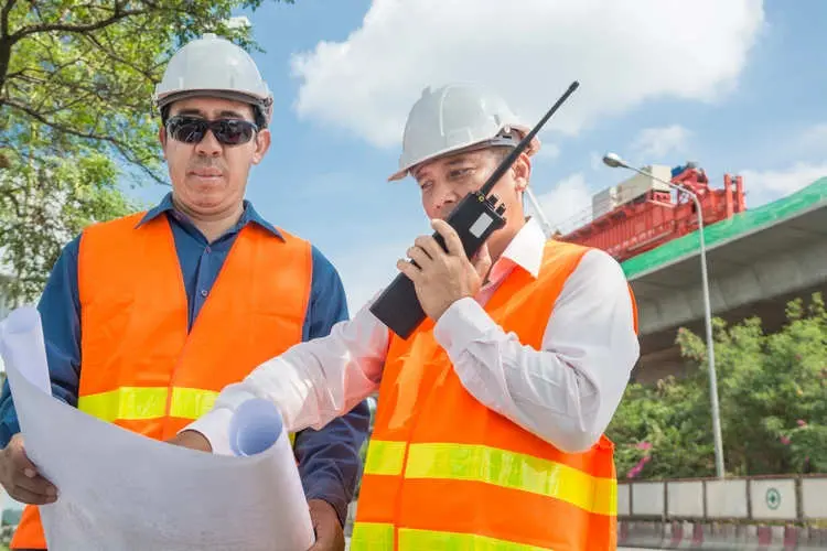 İş güvenliği uzmanları şantiyede proje inceliyor: baret, reflektif yelek ve walkie-talkie