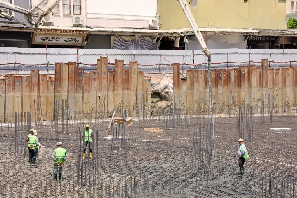 Kentsel inşaat sahasında geniş ölçekli temel imalatı: düz zemin üzerinde donatı hasırı, çok sayıda kolon filizi ve beton pompasıyla dökme işlemi