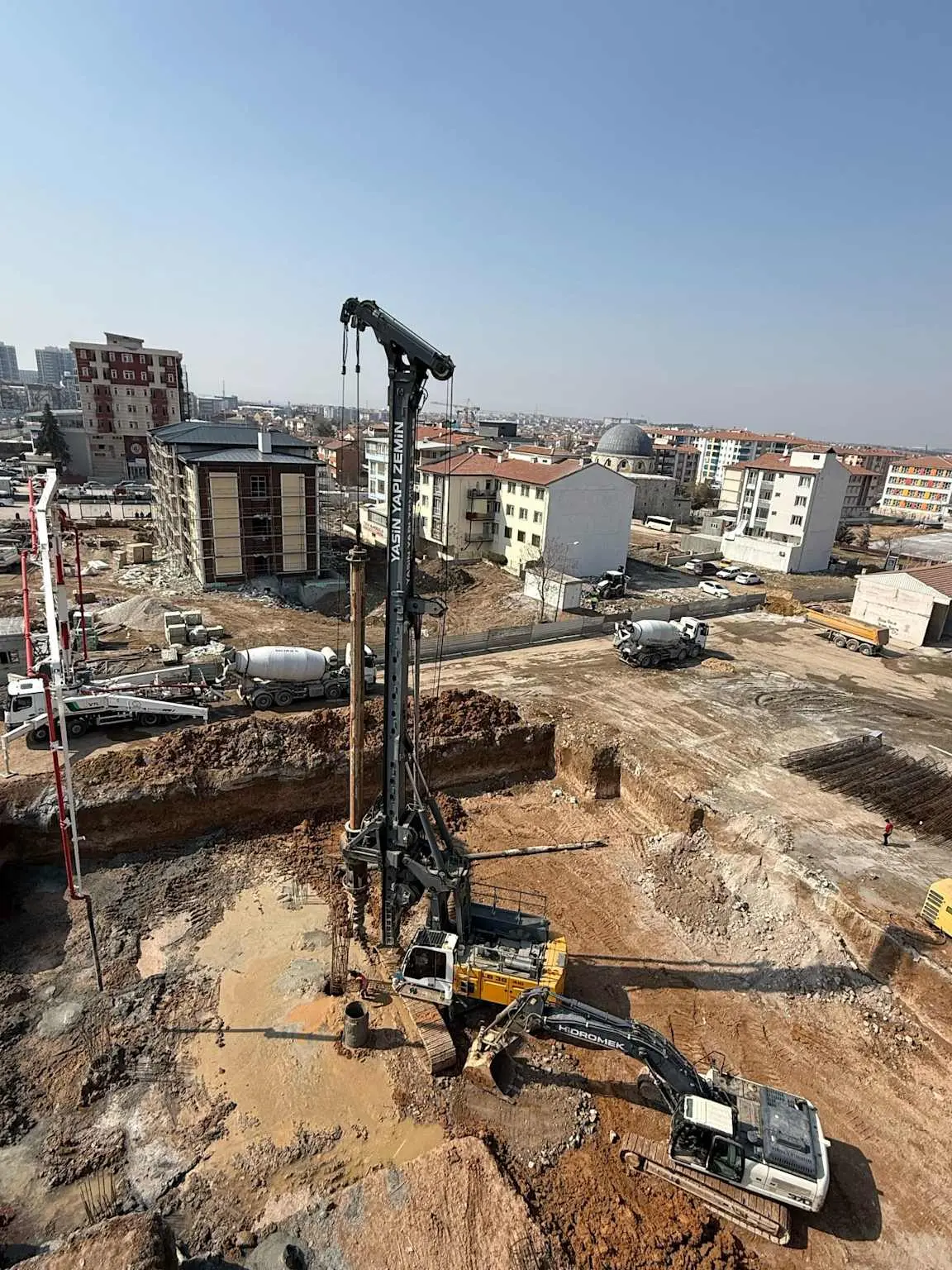 Türkiye'de şehir içi bir inşaat şantiyesinde fore kazık delgi makinesi çalışıyor; büyük çaplı sarı renkli delgi kulesi bir yapı bodrum kazısında konumlanmış, arka planda apartman binaları ve camii görülmektedir
