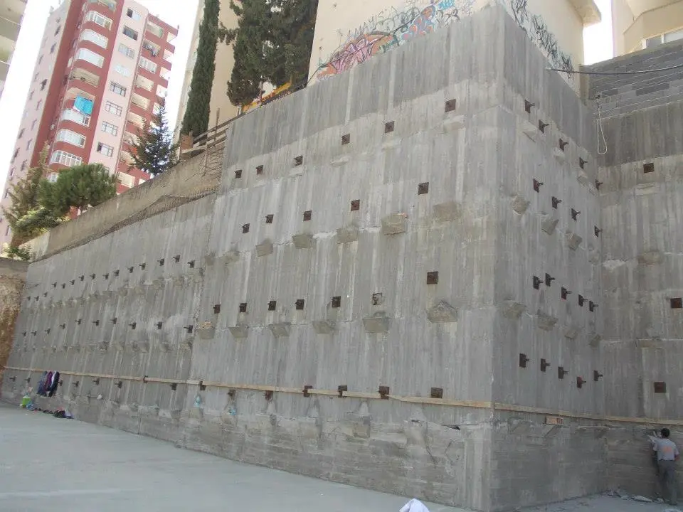 Kentsel alanda tamamlanmış büyük bir betonarme ankrajlı istinat duvarı: satır satır düzenli grid şeklinde ankaj başlıkları görünüyor, arka planda çok katlı bina
