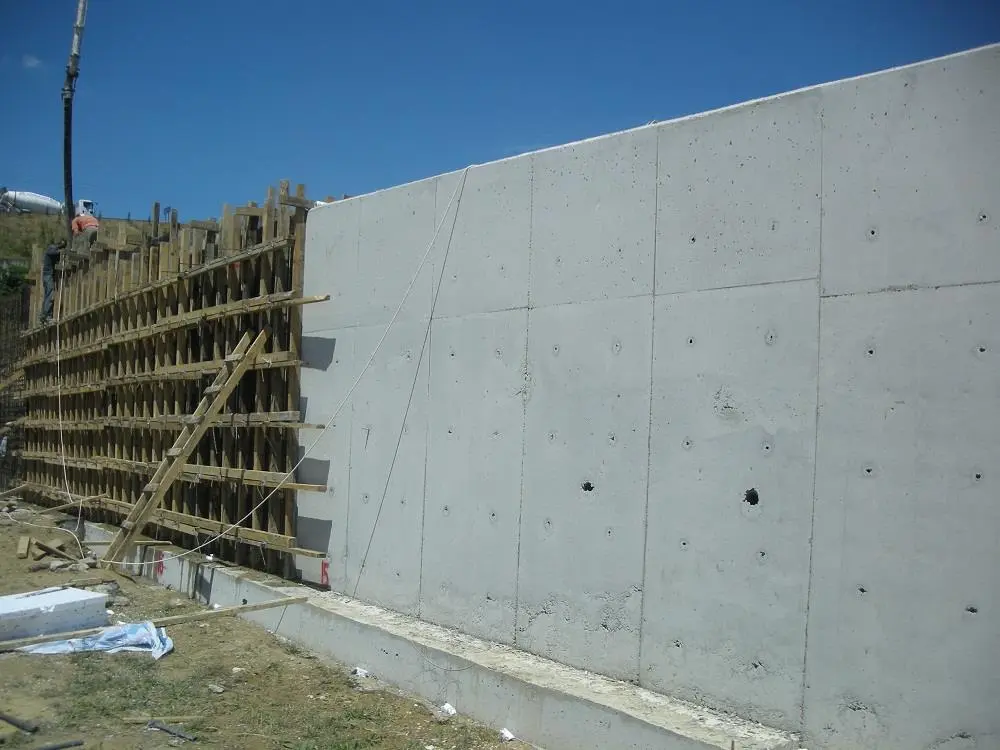 Tamamlanmış BA konsol istinat duvarı ön yüz görünüşü: düz beton yüzey, kalıp delik izleri, sol tarafta kaldırılan ahşap kalıp sisteminin bir kısmı, mavi gökyüzü