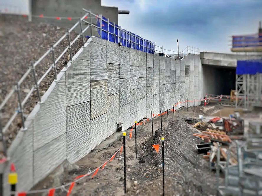 Türkiye şantiyesinde MSE toprakarme duvarı inşaatı: ön yüzde prefabrik beton paneller monte edilmiş, üst kısımda iskele ve mavi çelik yapı, ön zeminde kazı alanı ve bariyer konileri
