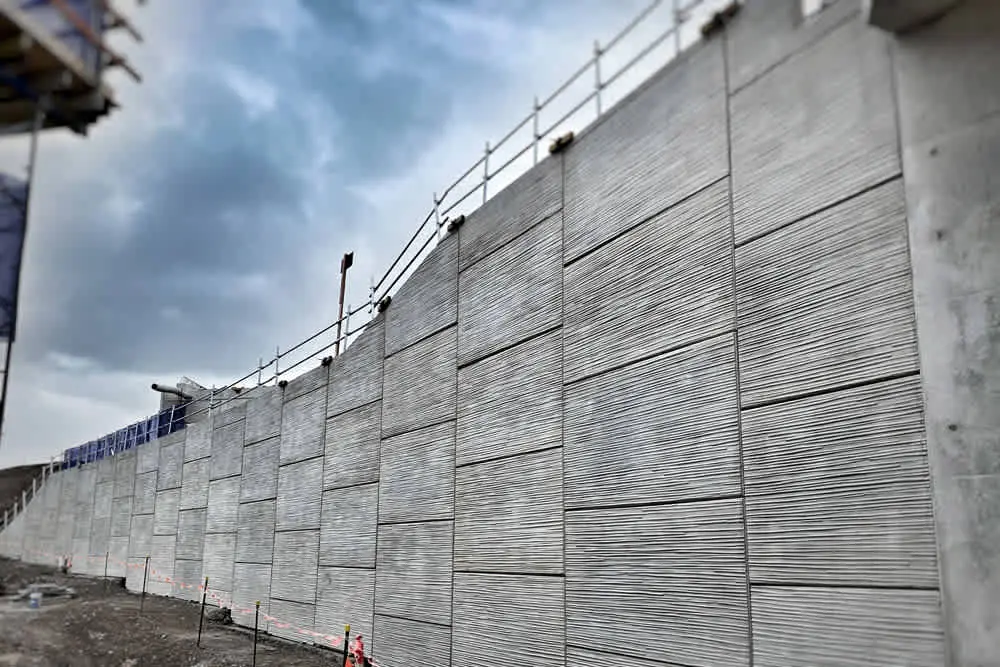 MSE toprakarme duvarı geniş panel ön yüzü yakın çekim: dikey çizgili yatay bantlı geniş beton prefabrik paneller; arka planda mavi bayrak ve bulutlu gökyüzü