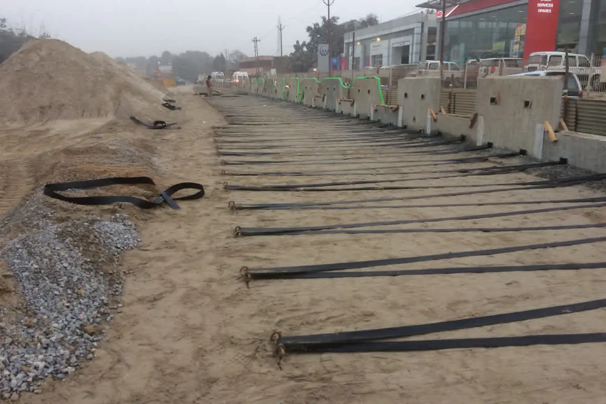 MSE duvarı inşaatında geogrid ve çelik şerit döşeme: taban dolgusu üzerine panel sırası önünde paralel uzanan koyu renkli geogrid şeritler/çelik çubuklar; arka planda kentsel ortam