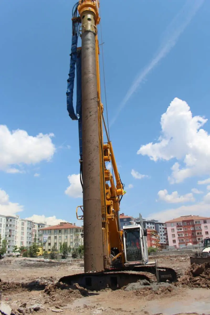 Büyük çaplı fore kazık makinesi — sarı paletli kazık delme ekipmanı, mavi gökyüzü, kentsel arka plan; Iç Anadolu'da konut inşaatı şantiyesi