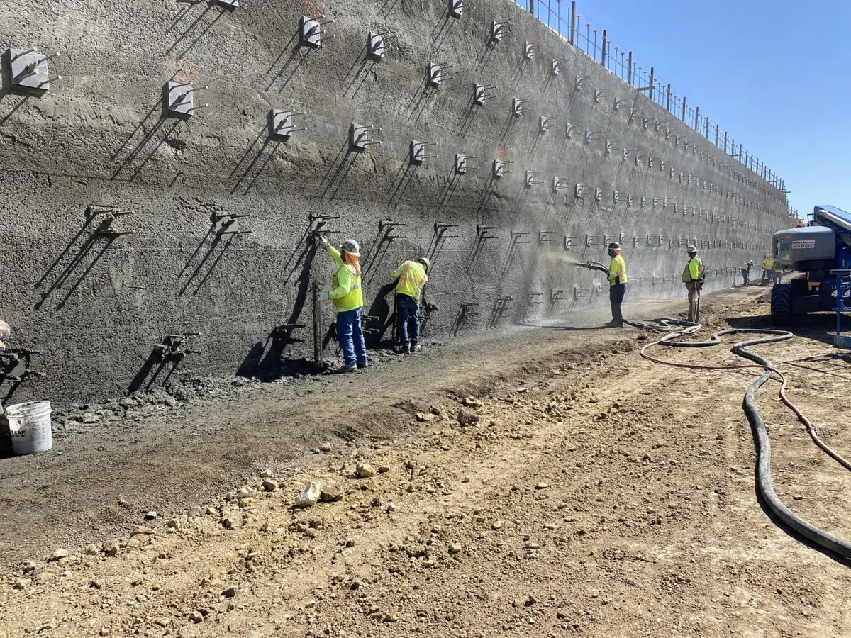 Zemin çivisi shotcrete duvar uygulaması — üç mühendis sarı yelekle dik kazı yüzeyine shotcrete spreyi uyguluyor, çivi başlık plakaları ve hasır donatı gözüküyor