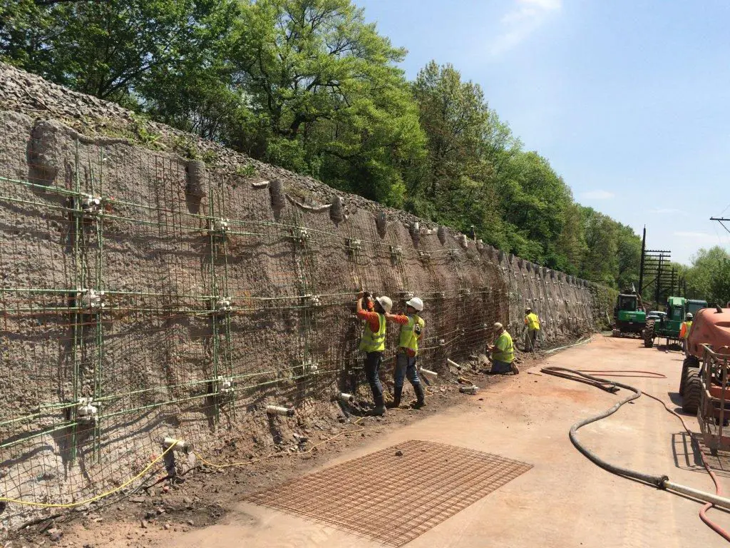 Yol yarması üzerinde zemin çivisi hasır donatı uygulaması — yeşil ağaçlı yol kenarında yüksek dik kazı yüzeyine hasır donatı ve çivi başlıkları yerleştirilmiş, shotcrete öncesi aşama