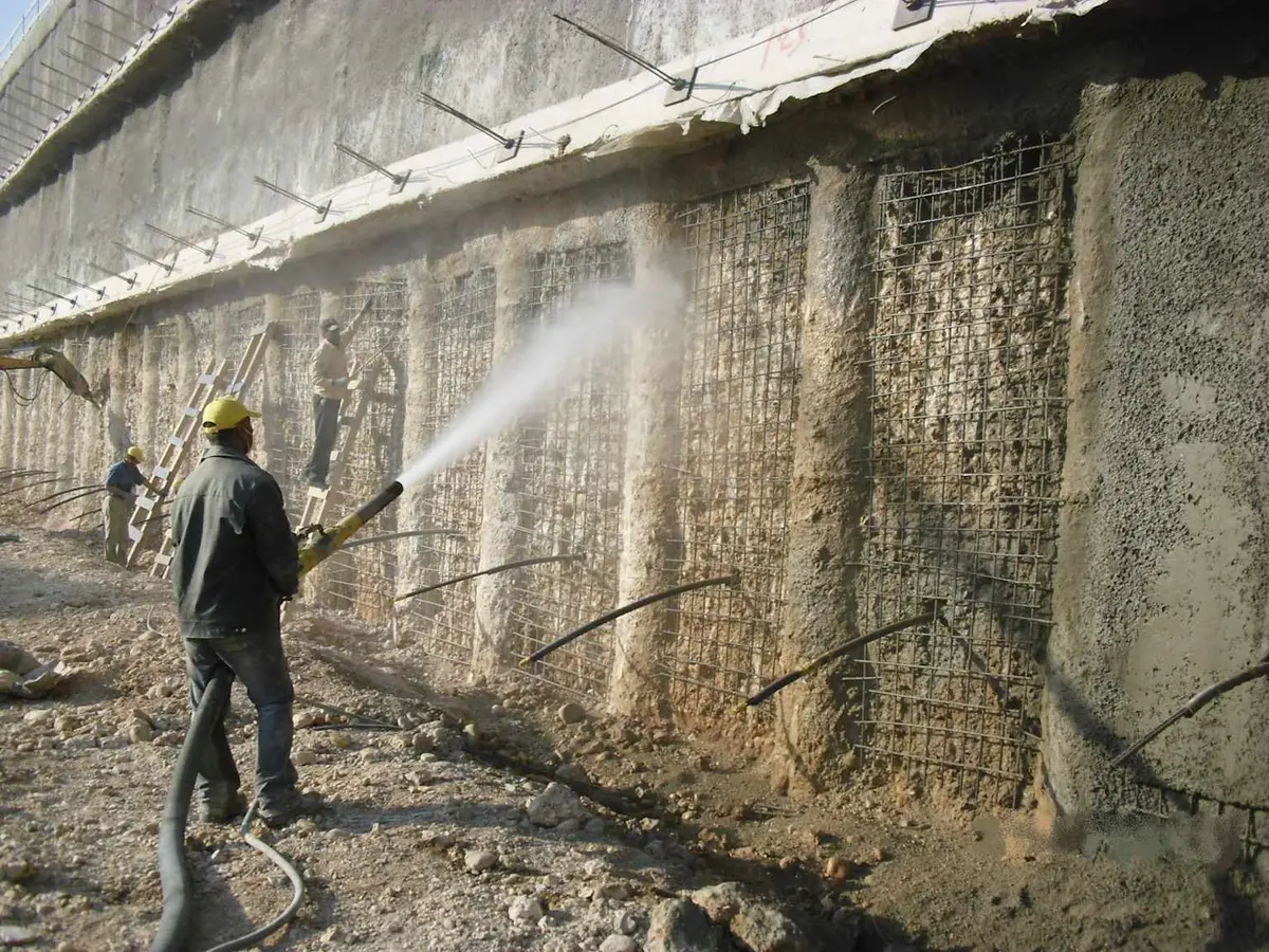 İşçi kazı yüzeyine shotcrete uygulaması yapıyor — uzun hortum ile püskürtme beton uygulaması, yüzeyde çivi başlıkları ve hasır donatı görünüyor, arka planda beton köprü