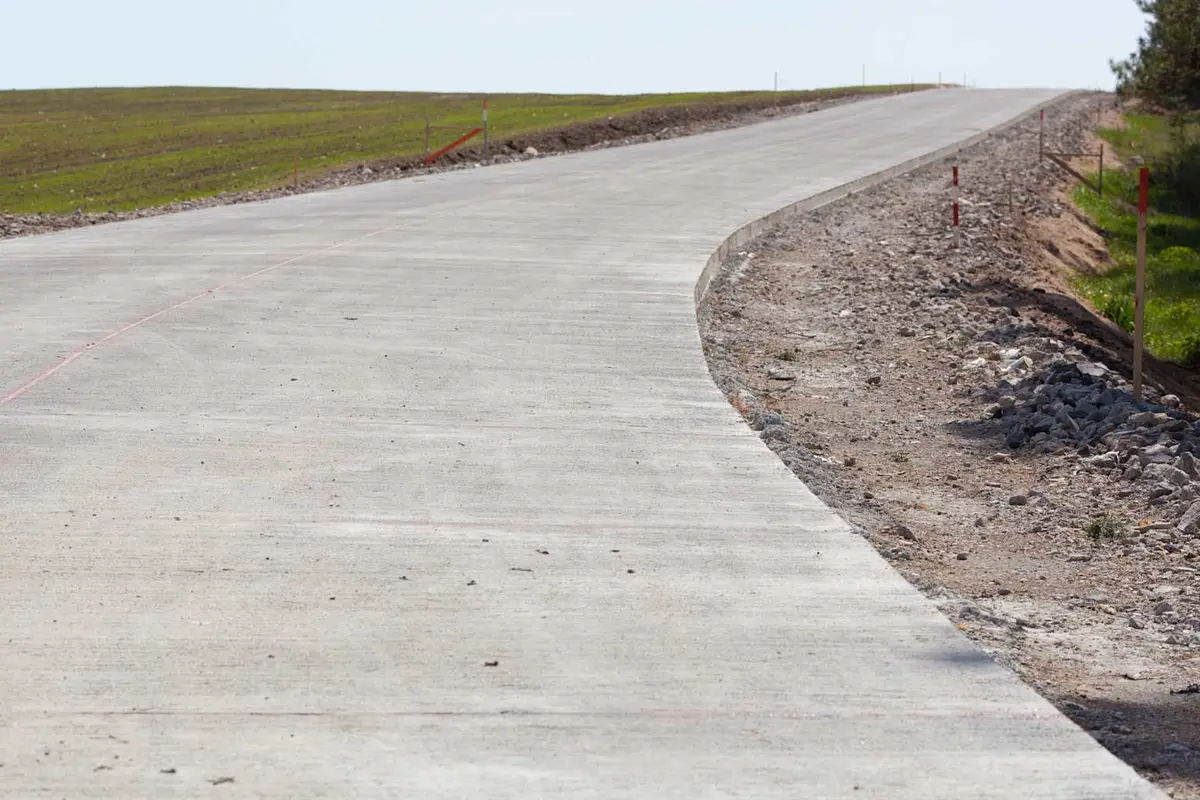 Kırsal kesimde taze dökülmüş JPCP beton yol kaplama uygulaması: beton kaplama yüzeyi pürüzlü tekstür; yolun sol kenarında granüler zemin hazırlığı ve kırmızı şerit kazıklar; arka planda yeşil çimen ve ağaçlık alan; hafif kavisli yol ekseni