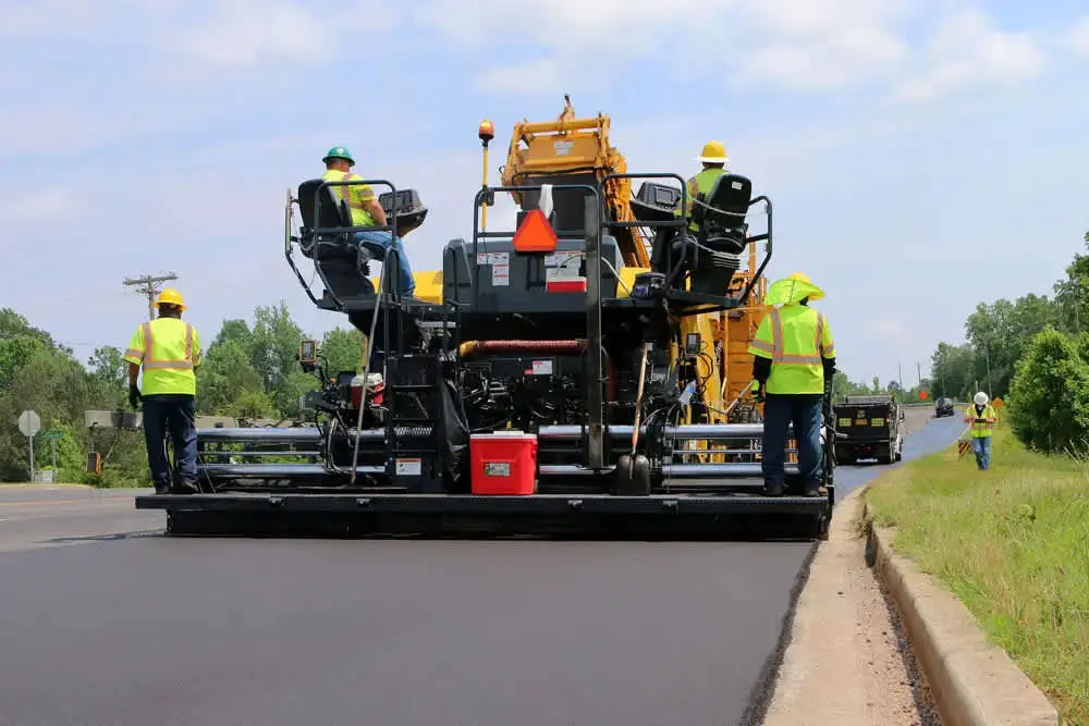 BSK asfalt serme makinesi (finisher) ile yol inşaatı: sarı Caterpillar finisher asfalt serme yaparken; üç işçi yeşil fosforlu yeleklerle çalışmakta; sol tarafta bitişik beton bordür; bitki örtüsü ve hafif bulutlu gökyüzü arka planda görülmektedir