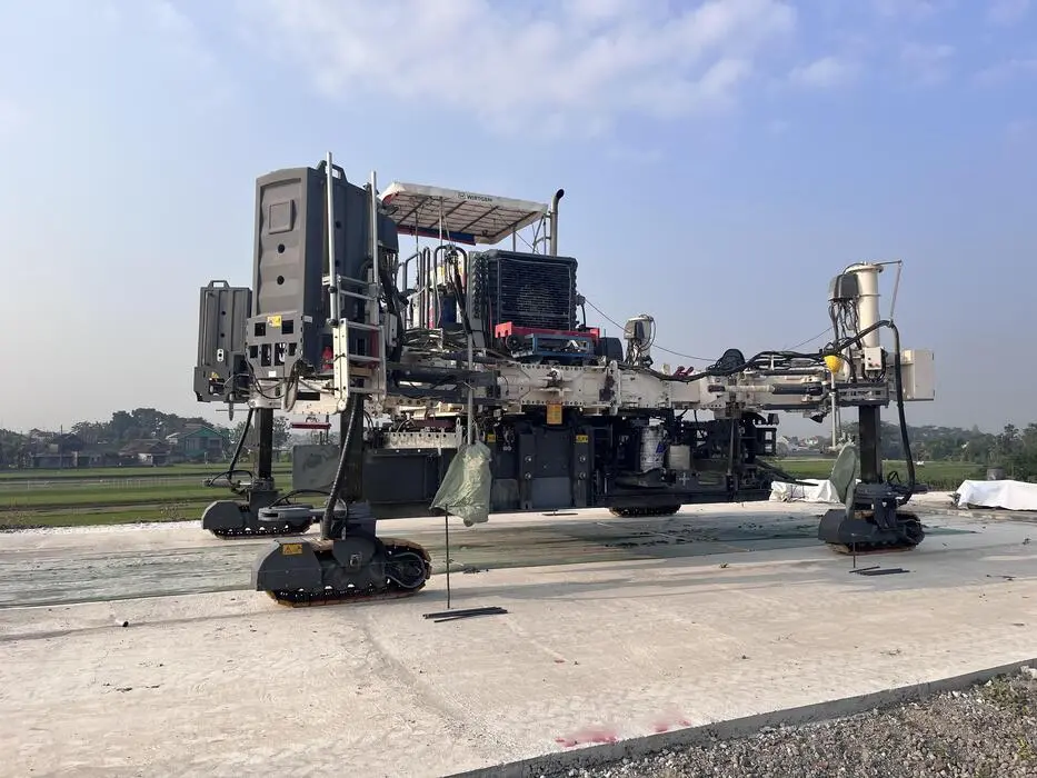 Açık alanda çalışan büyük bir kayar kalıp (slipform paver) makinesi; beton yol döküm işlemi sırasında makine geniş şerit üzerinde beton yayıyor ve sıkıştırıyor