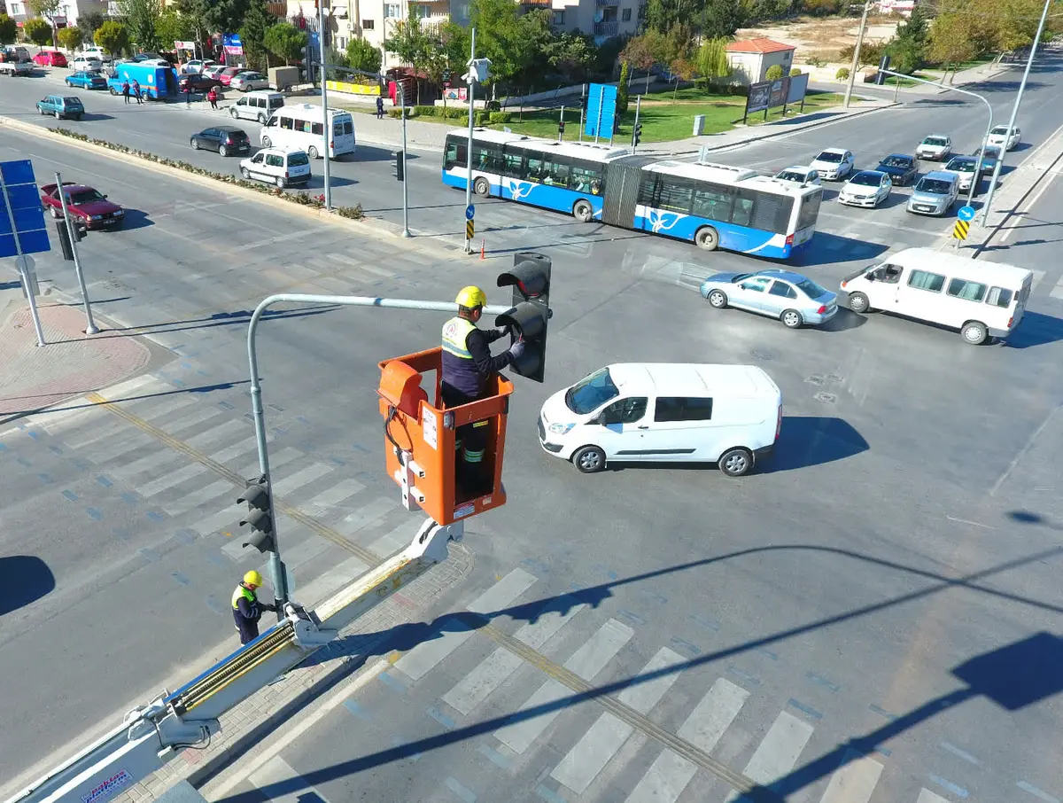 Türkiye'de bir şehirde sinyalize kavşakta trafik sinyali direğinde yüksek menevreli platformda çalışan iki işçi; altta kavşaktan geçen araçlar ve servis otobüsleri görülüyor