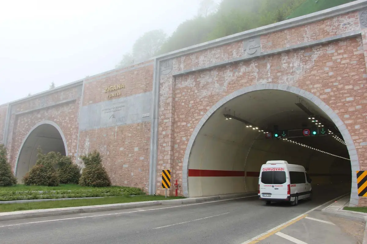 Sisli dağ ortamında kırmızı kiremit desen kaplamali çift portallı Zigana Tüneli girişi; sol portaldan ambulans çıkıyor, sağ portal karanlık; portal çevresinde ağaçlar ve sis