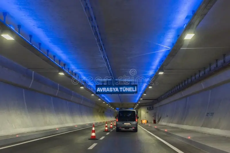 Avrasya Tüneli iç bölgesi; iki şeritli yüksek tavanlı tünel, mavi LED vurgu aydınlatması tavan kenarlarında, LED panel dizileri tavanda, ortada AVRASYA TÜNELİ yazılı mavi LED tabela, öndeki araç