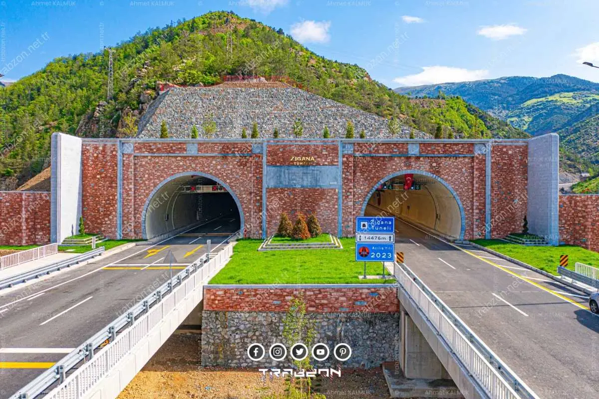 Dağlık yeşil arazi önünde kırmızı taş kaplama portal duvarlarıyla çerçevelenmiş iki tüplü karayolu tünel girişi; iki tüp arasında yeşil refüj bandı
