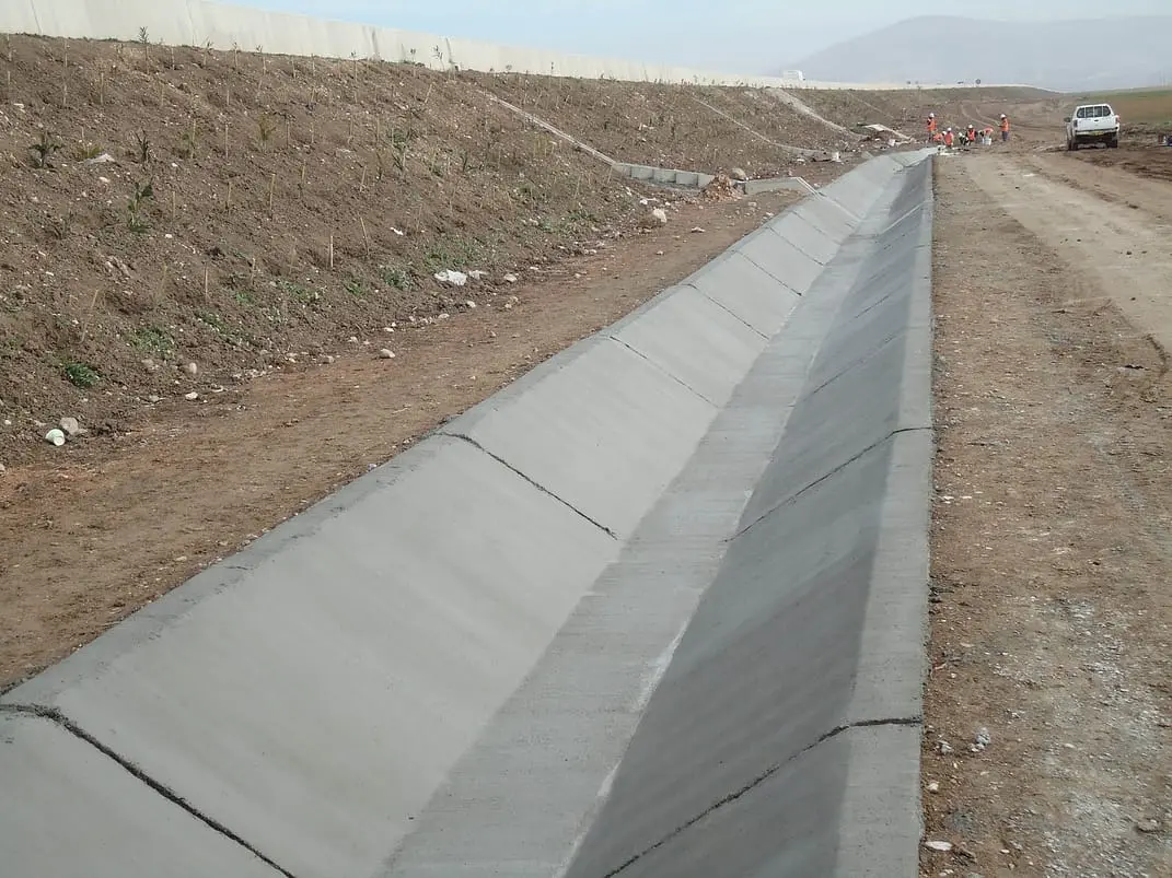 İnşaat aşamasında beton kaplı trapez yol hendeği; solda toprak şev, sağda çakıl zemin; uzun boyuna hendek kesiti; arkada işçiler ve beyaz beton duvar