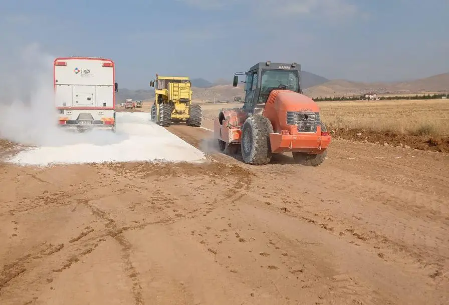 İç Anadolu kuru step arazisinde karayolu inşaatında kireç stabilizasyonu: beyaz kireç serpme makinesi zemine kireç serperken arkasında sarı greyder ve turuncu silindir görülmektedir
