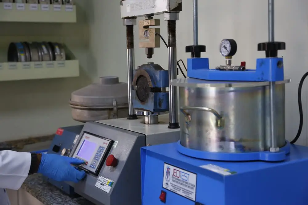 Mavi gövdeli JEOTEST markalı Marshall stabilite test cihazı: yük uygulama kafası, basınç manometresi ve su banyosu ile test laboratuvarında çalışan teknisyen