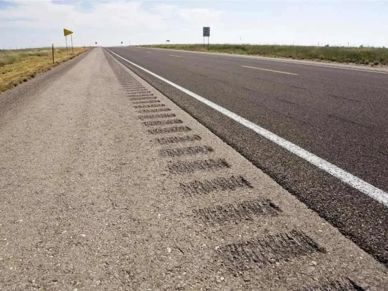 Tırmanma şeridi (climbing lane) uygulaması: yatay düz asfalt yolun sağ kenarında rumble strip (ses şeridi) ve genişletilmiş banket alanı — uzak perspektifte sarı uyarı işareti görülüyor